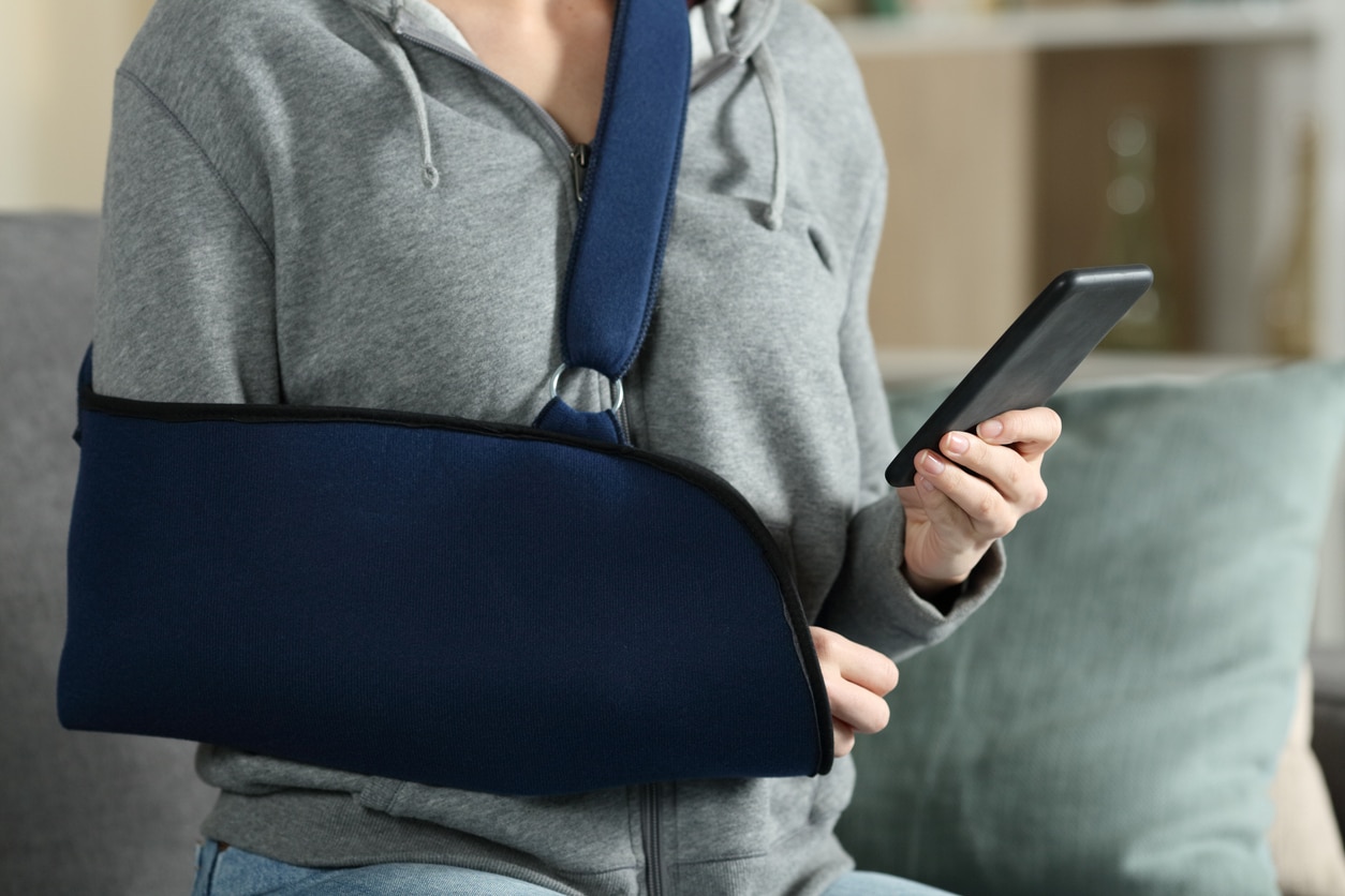 Close up of woman hands with broken arm wearing sling using smart phone at home